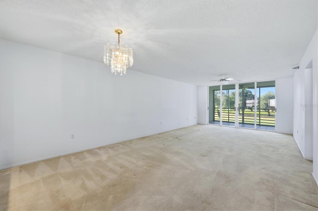 An empty living room with beige carpet, a crystal chandelier, and sliding glass balcony doors — 3740 PINEBROOK CIRCLE UNIT 207, BRADENTON, FL