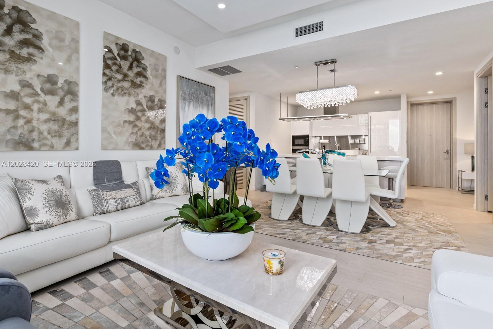 A bright living and dining area features white furniture, a crystal chandelier, and blue orchids — 1000 Brickell Plz, Miami FL 33131, MIAMI, FL