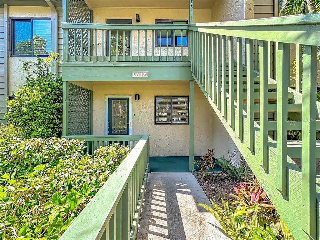 A two-story apartment building exterior features green wooden railings, stairs, and a ground-level entrance — 1515 PELICAN POINT DRIVE UNIT BA185, SARASOTA, FL
