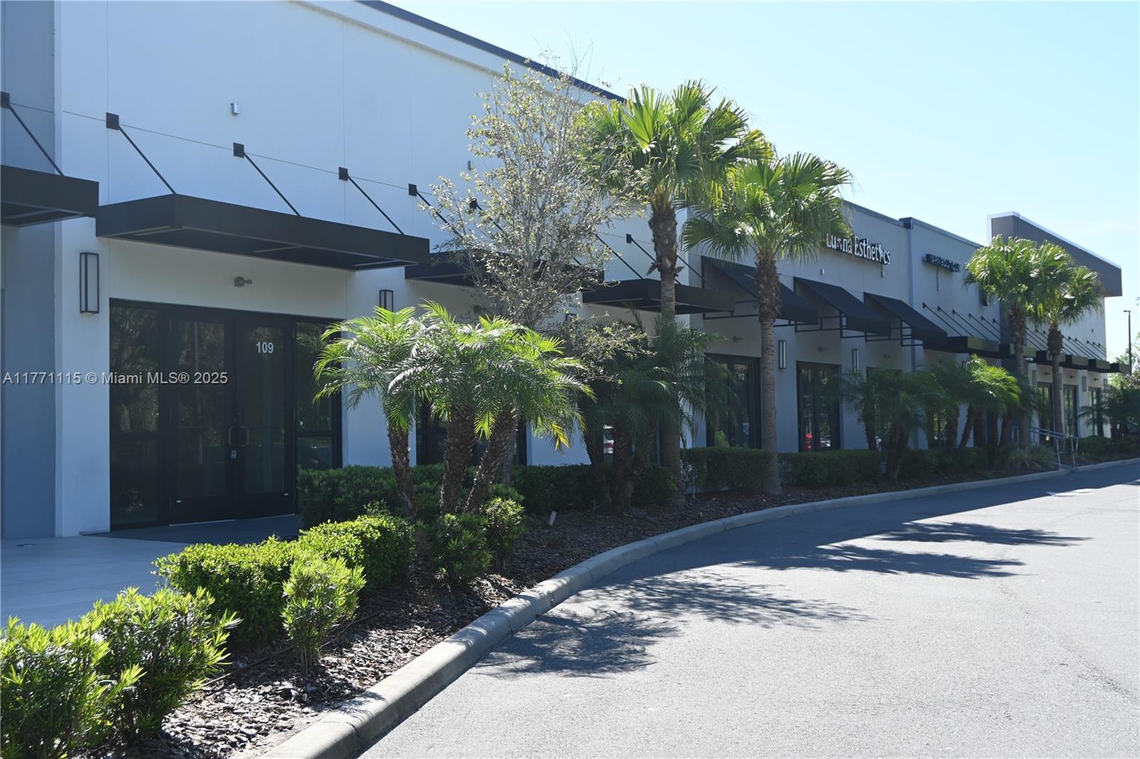 A modern commercial building exterior features glass storefronts, black metal awnings, and landscaped palm trees — 3370 ROUSE RD # 109, Orlando FL 32817, ORLANDO, FL