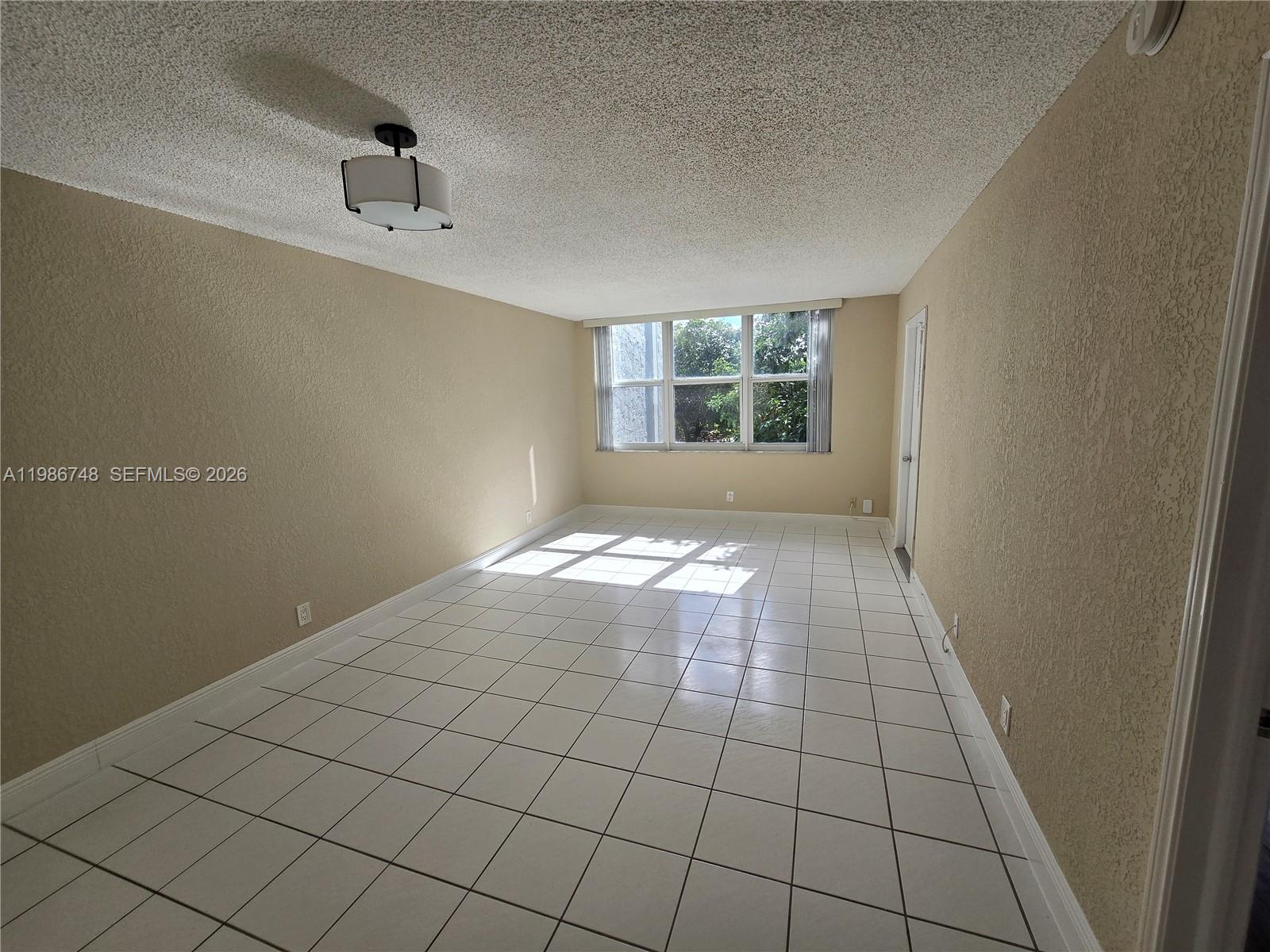 An empty room features white tile flooring, beige walls, a popcorn ceiling, and a window — 9461 Evergreen Pl # 305, Davie FL 33324, DAVIE, FL