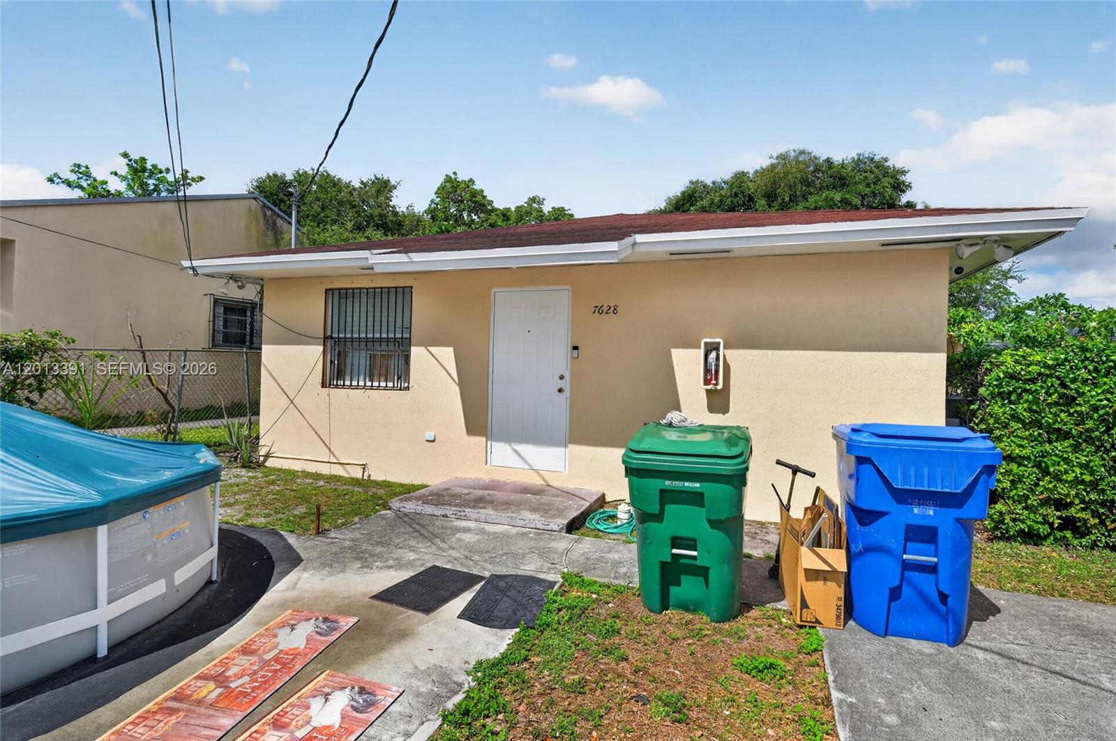 A single-story house exterior features a white door, a window with bars, and two trash bins — 7628 NW 2nd Ct, Miami FL 33150, MIAMI, FL