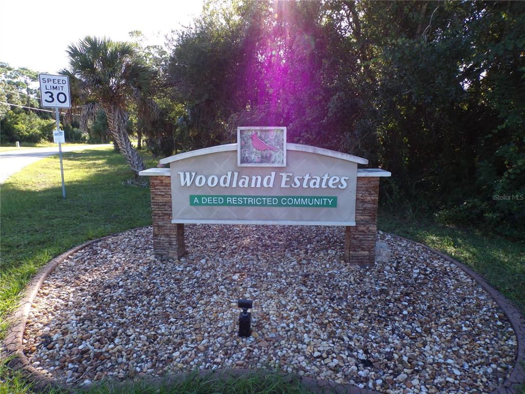 A stone monument sign for Woodland Estates stands in a gravel bed near a road — 25079 HARBORSIDE BOULEVARD, PUNTA GORDA, FL