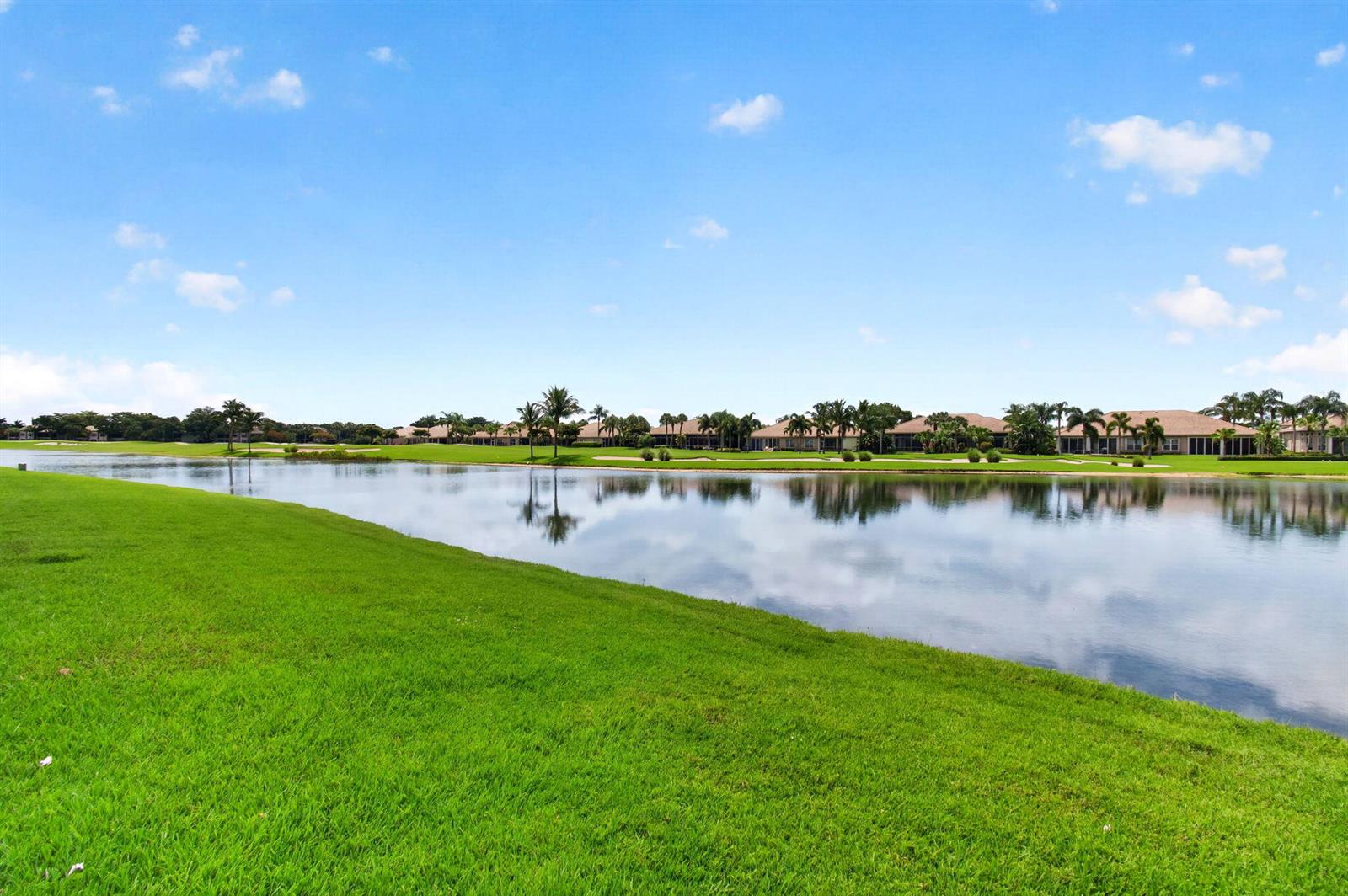A grassy lawn overlooks a lake with houses in the background under a blue sky — 7004 Bitterbush Place, Boynton Beach FL 33472, BOYNTON BEACH, FL