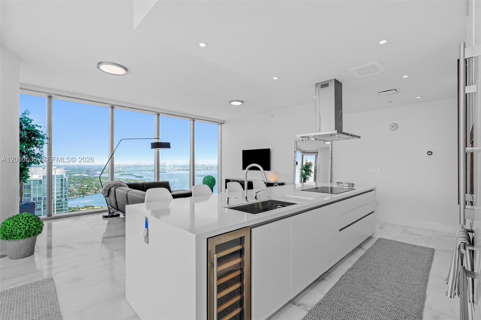 A modern white kitchen island with a wine cooler overlooks a living area and bay — 700 NE 26th Terrace # 5606, Miami FL 33137, MIAMI, FL