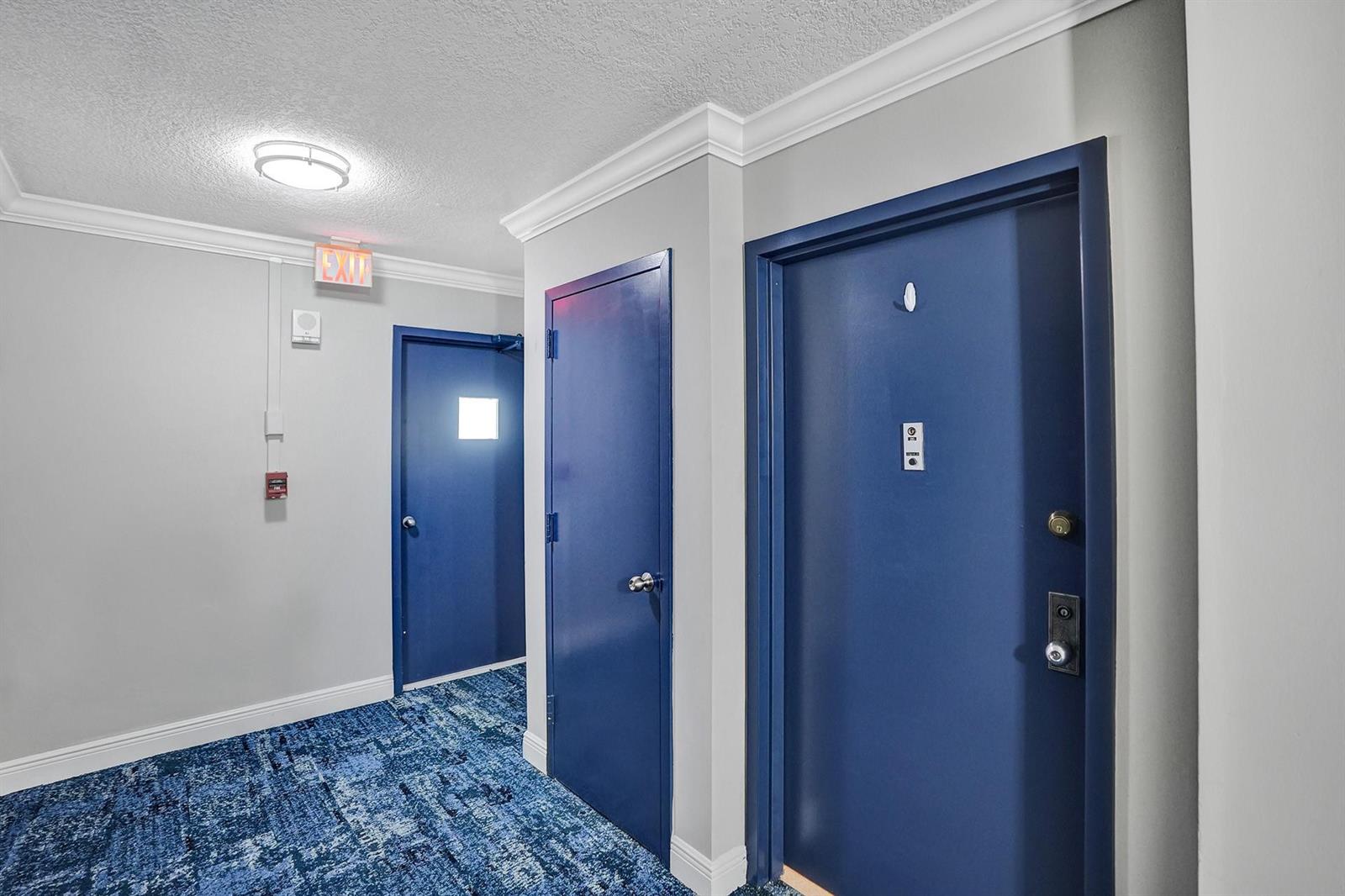 A hallway with blue doors, gray walls, and blue patterned carpeting leads to an exit — 1361 S Ocean Bl # 210, Pompano Beach FL 33062, POMPANO BEACH, FL