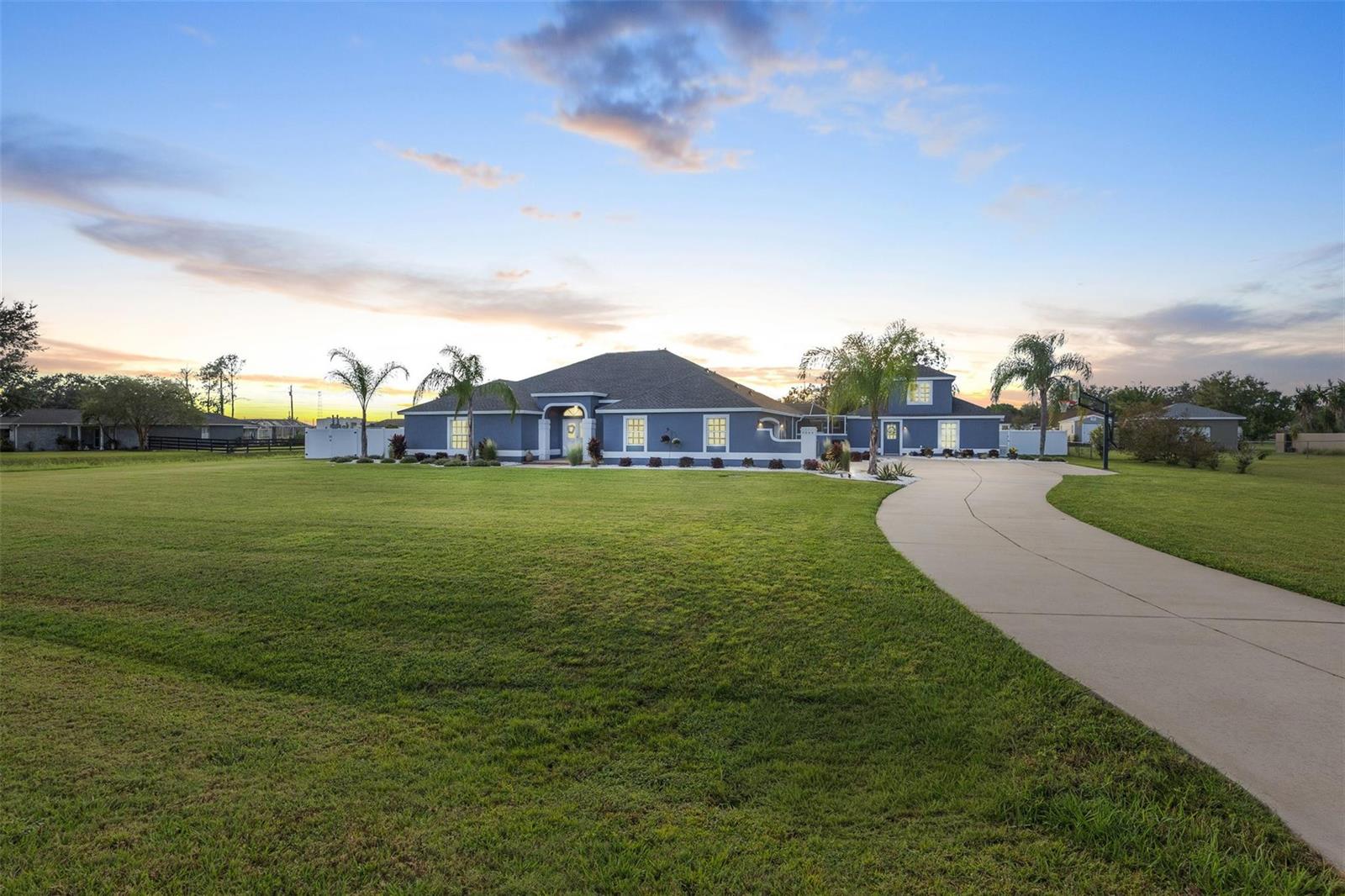 A blue single-story house with a large lawn and a curved concrete driveway at sunset — 9060 County Road 128c, Wildwood FL 34785, WILDWOOD, FL
