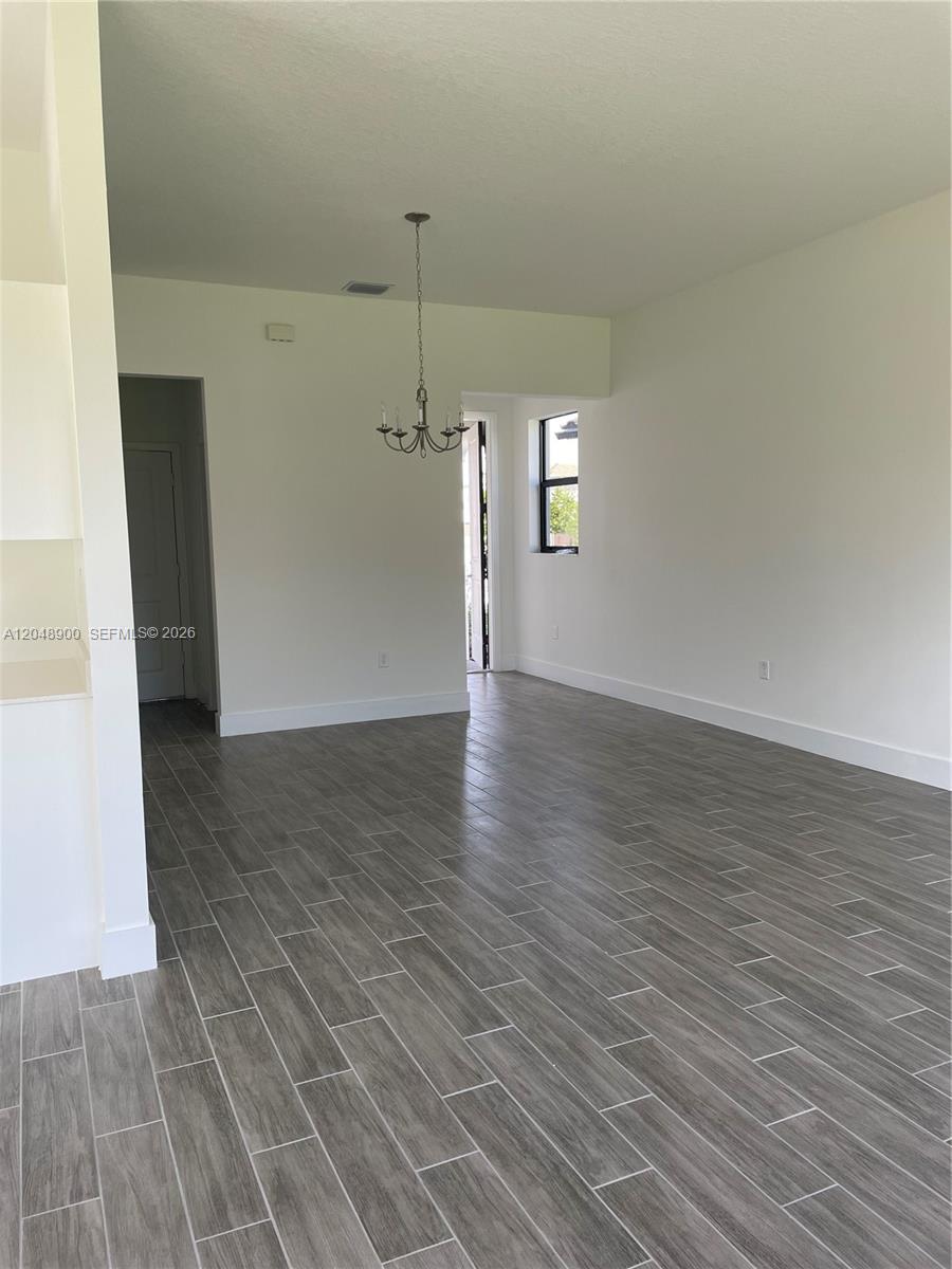 An empty room features gray wood-look tile flooring, white walls, and a hanging chandelier — 5496 Agostino Way, Ave Maria FL 34142, AVE MARIA, FL