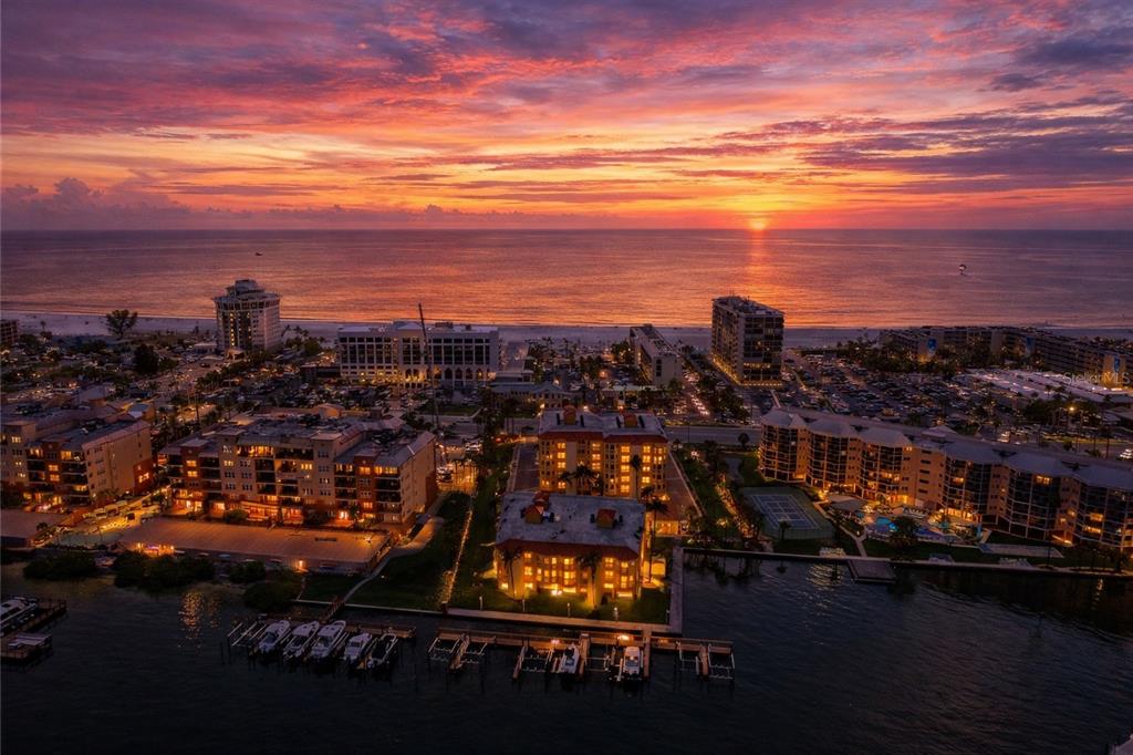 An aerial view shows a coastal town with multi-story buildings, a beach, and a sunset — 5353 GULF BOULEVARD UNIT A404, ST PETE BEACH, FL