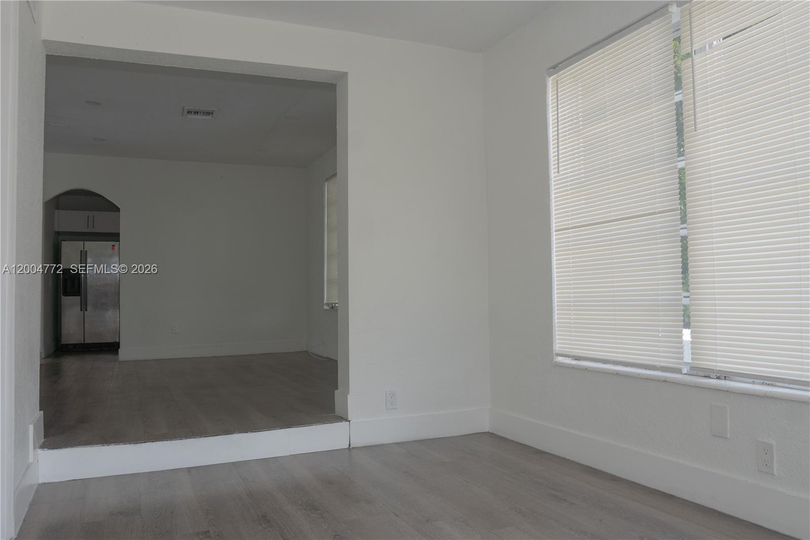 A room with light wood-look flooring and a large window leads to a kitchen area — 3376 NW 49th St, Miami FL 33142, MIAMI, FL