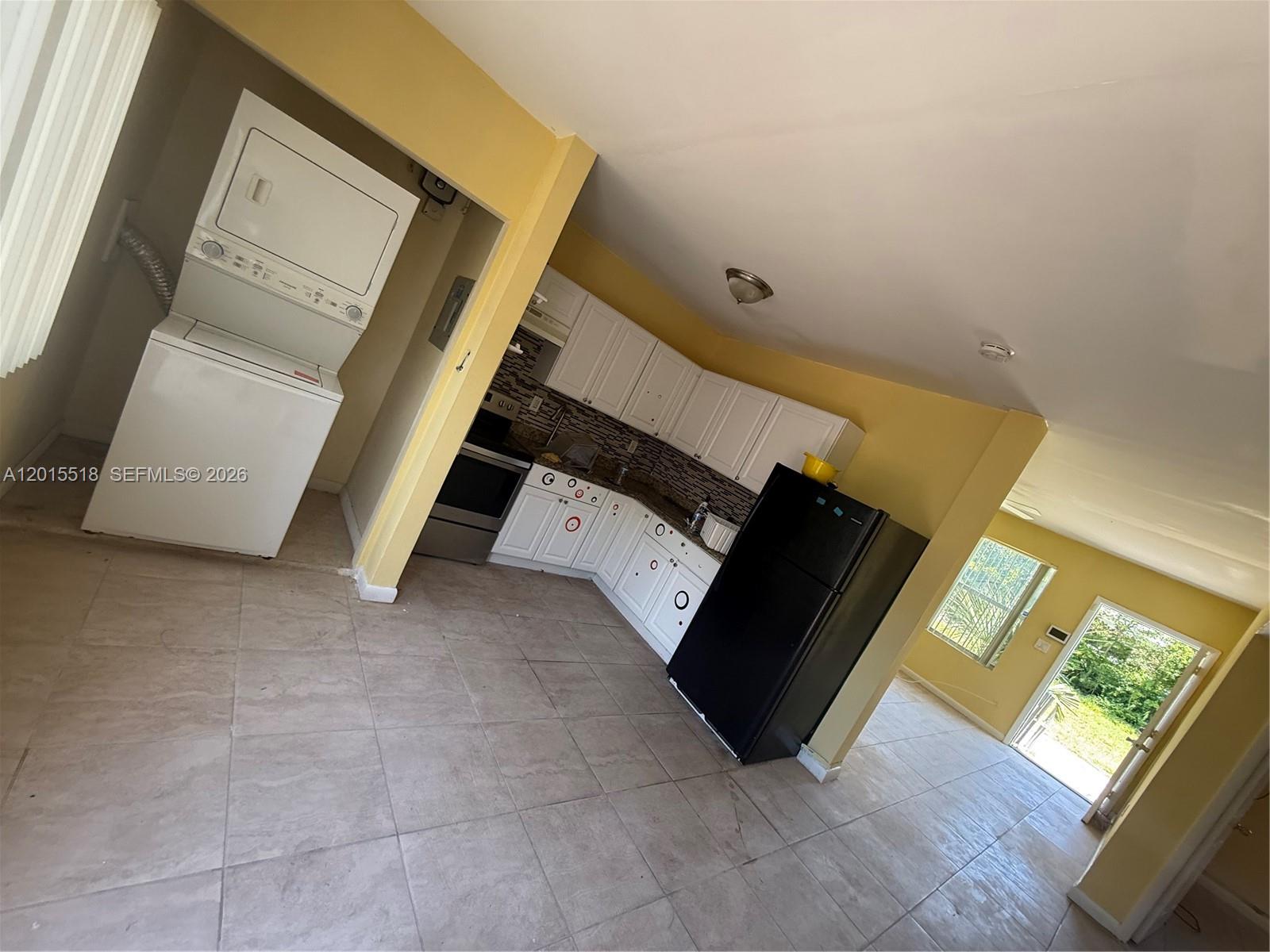 This kitchen features white cabinets, a black refrigerator, a stacked washer-dryer, and tile flooring — NORTH MIAMI BEACH, FL