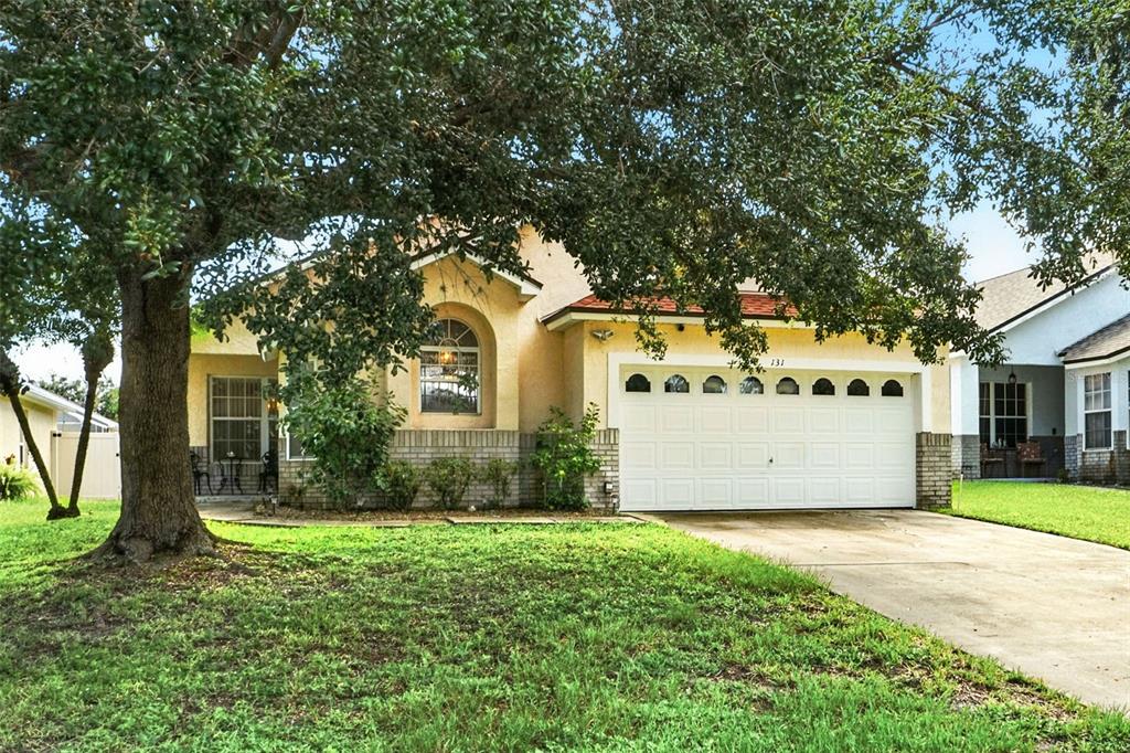 A single-story house with a white garage door, tan stucco walls, and a large front tree — 131 S ROMA WAY, KISSIMMEE, FL