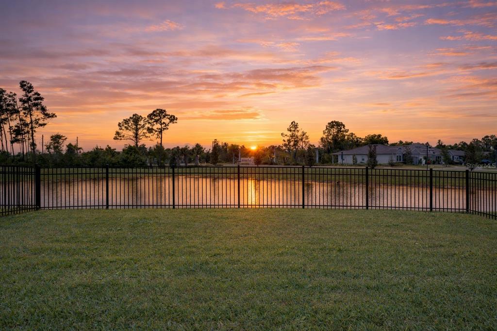 A fenced backyard overlooks a pond at sunset with trees and houses in the distance — 7024 LANIER FALLS ROAD, DELAND, FL