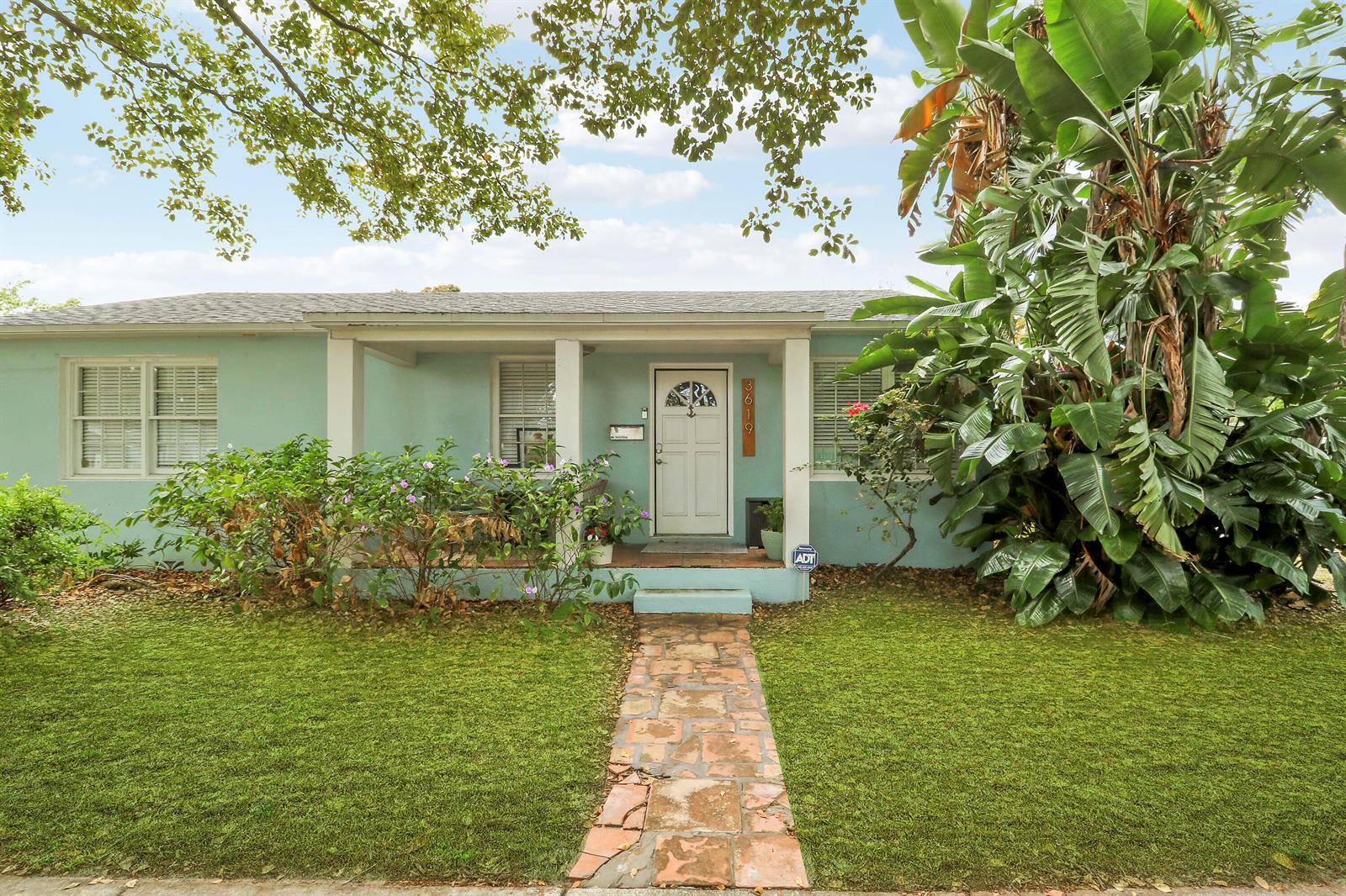A light blue single-story house features a front porch, stone walkway, and large tropical plants — 3619 Spruce Avenue, West Palm Beach FL 33407, WEST PALM BEACH, FL