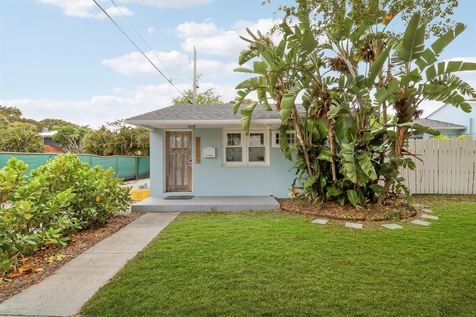 A light blue cottage with a wooden door and large tropical plants in the front yard — 3619 Spruce Avenue, West Palm Beach FL 33407, WEST PALM BEACH, FL