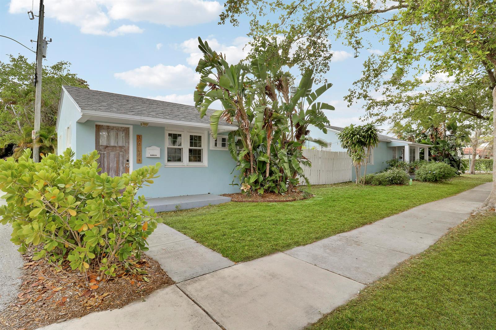 A light blue single-story house with a wooden front door and lush landscaping — 3619 Spruce Avenue, West Palm Beach FL 33407, WEST PALM BEACH, FL