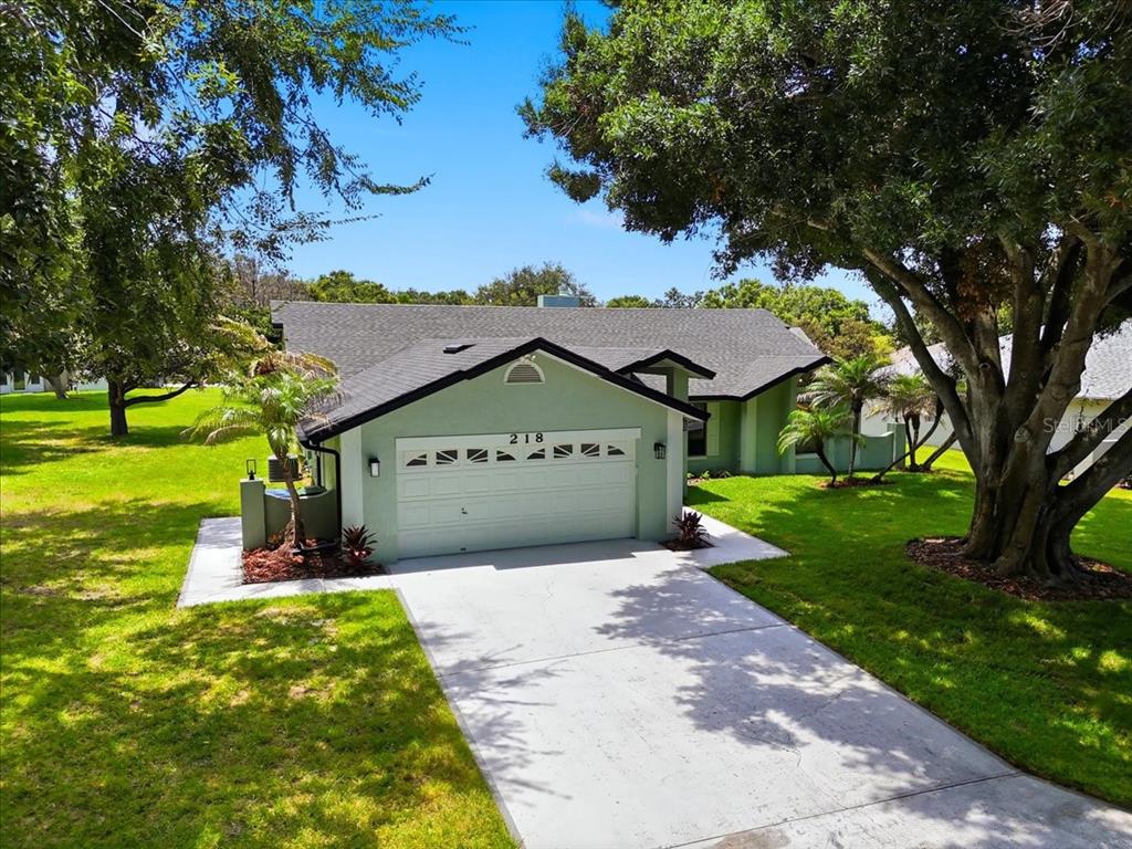A light green house with a two-car garage and a concrete driveway under large trees — 218 LAKE HARTRIDGE DRIVE NW, WINTER HAVEN, FL