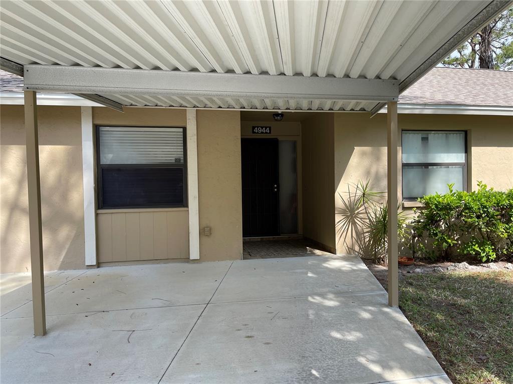 A beige house exterior features a concrete carport, a dark front door, and two windows — 4944 VILLAGE GARDENS DRIVE UNIT 151, SARASOTA, FL