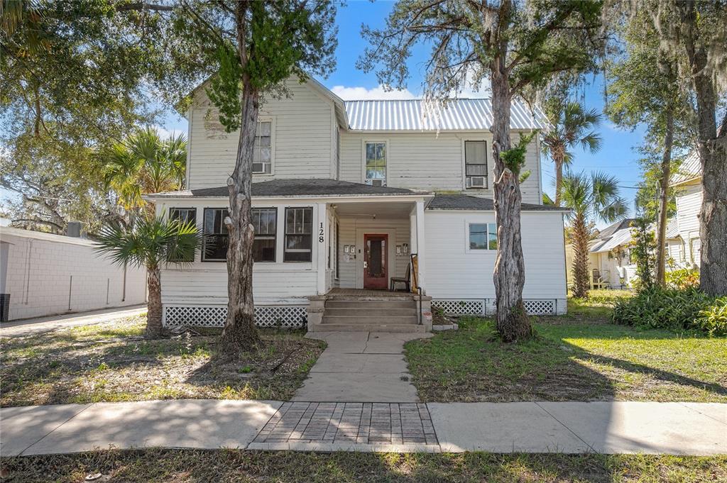 A two-story white house with a metal roof, front porch, and two large trees — 128 E RICH AVENUE, DELAND, FL