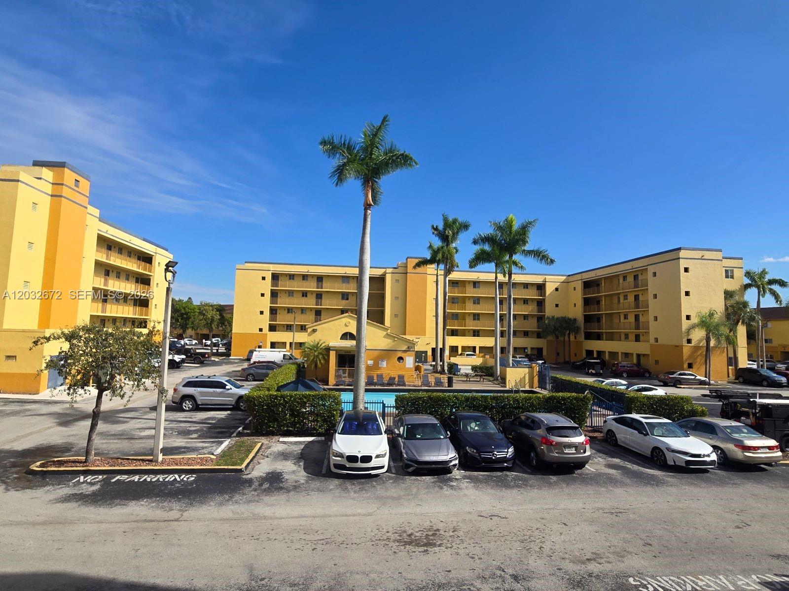 An exterior view of a yellow apartment complex with a parking lot and palm trees — 14961 SW 82nd Ln # 202, Miami FL 33193, MIAMI, FL