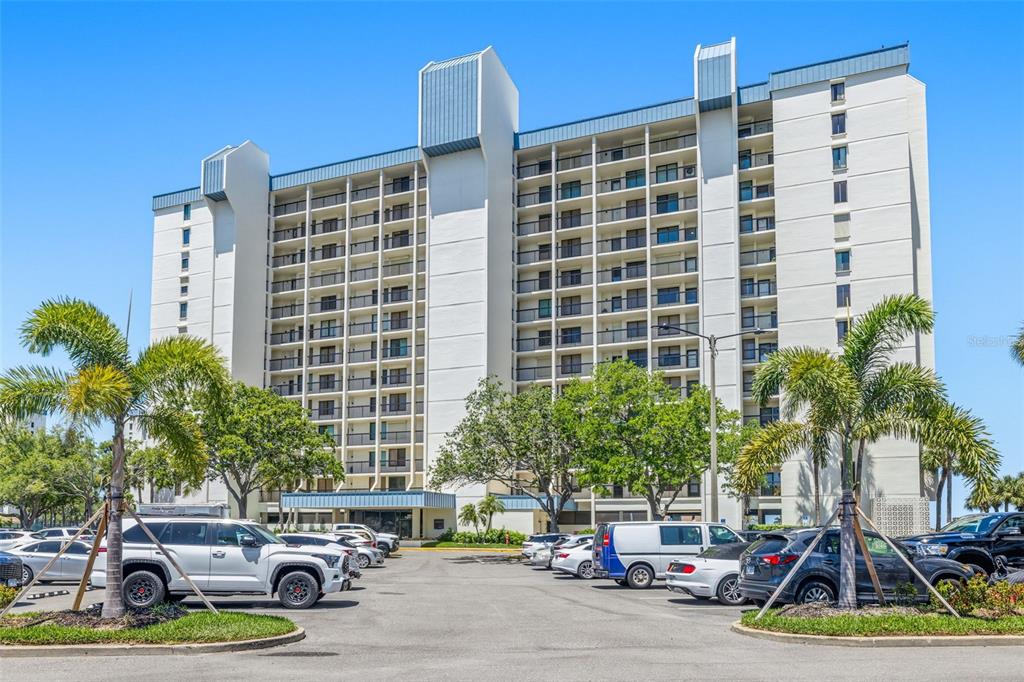 A multi-story residential building with balconies stands behind a parking lot filled with cars — 9425 BLIND PASS ROAD UNIT 705, ST PETE BEACH, FL
