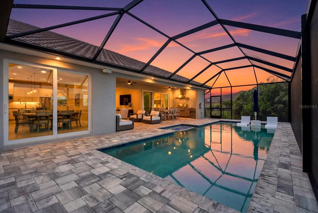 A screened-in pool patio features stone pavers, outdoor seating, and a view of the sunset — 15672 LAKELAND CIRCLE, PORT CHARLOTTE, FL