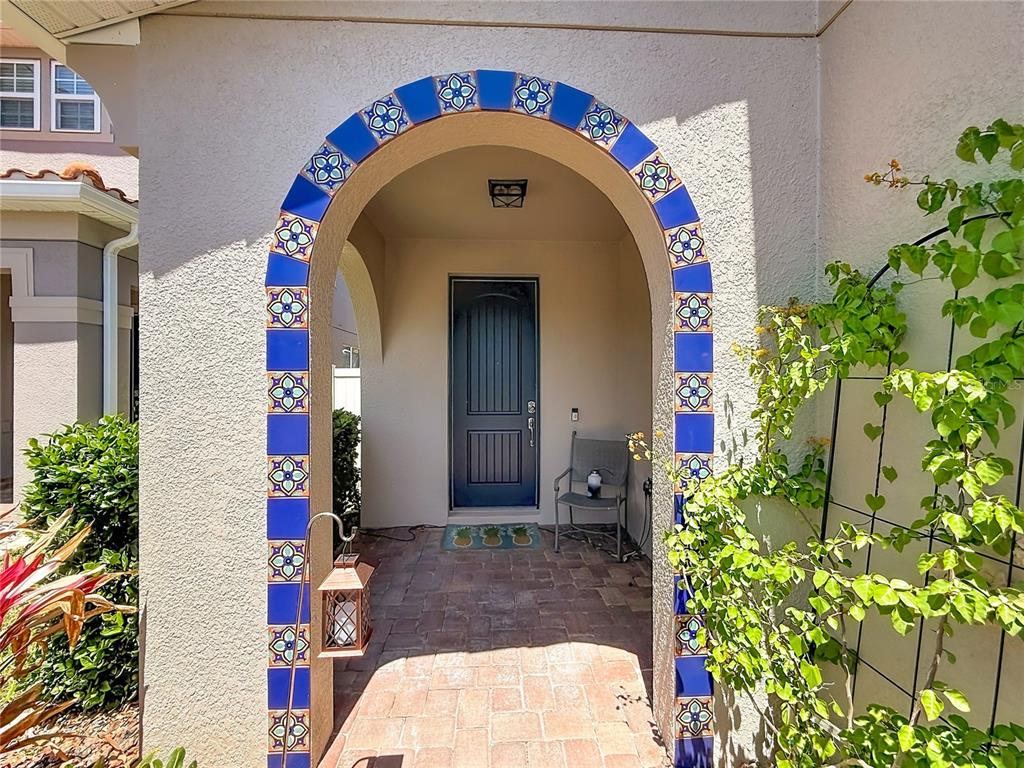 An arched entryway features blue decorative tiles and a dark door on a paved porch — 10193 GRAND OAK CIRCLE, MADEIRA BEACH, FL