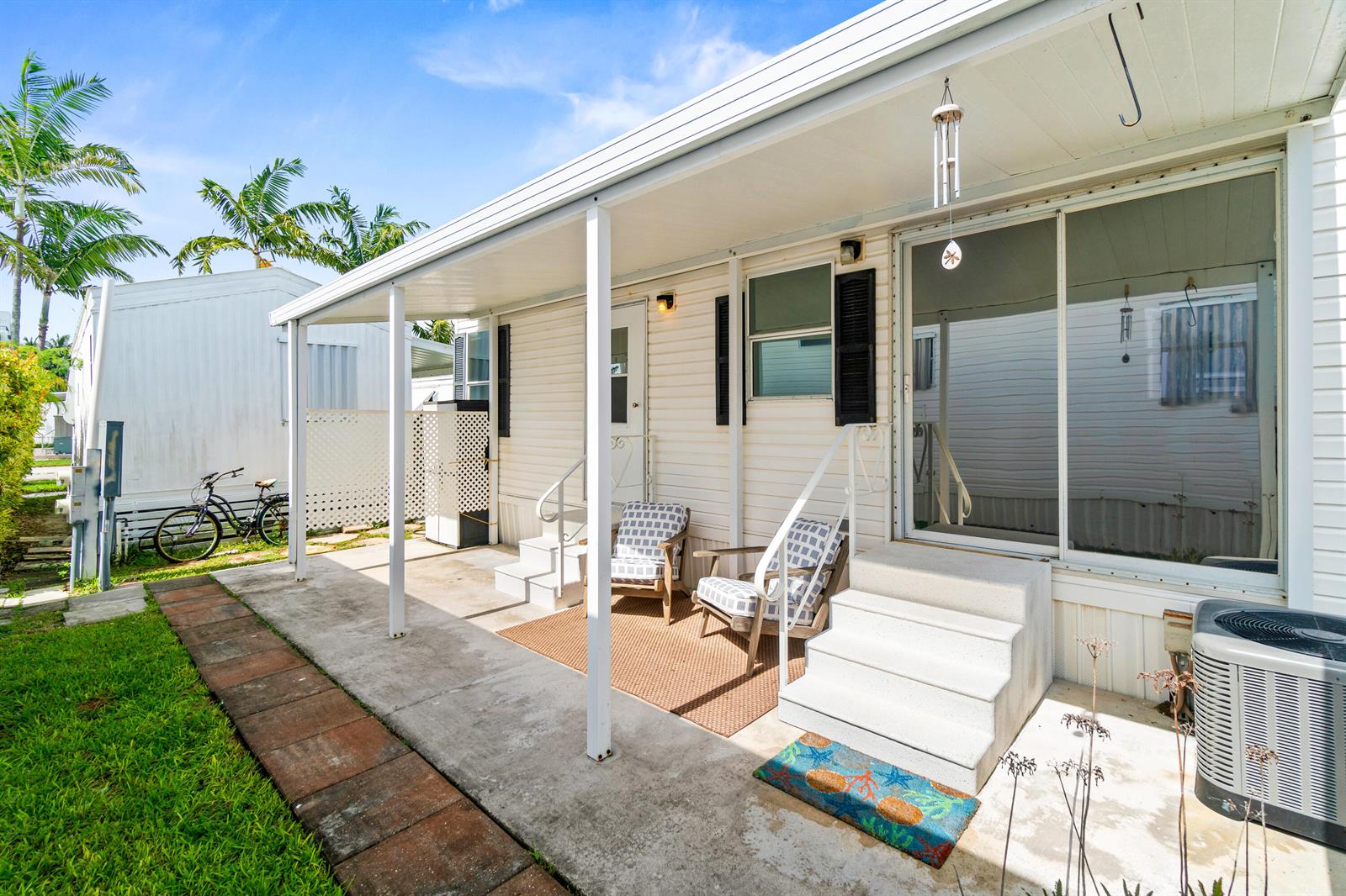 A mobile home exterior features a covered concrete patio with two chairs and white stairs — 12 Hibiscus Drive # H, Briny Breezes FL 33435, BRINY BREEZES, FL