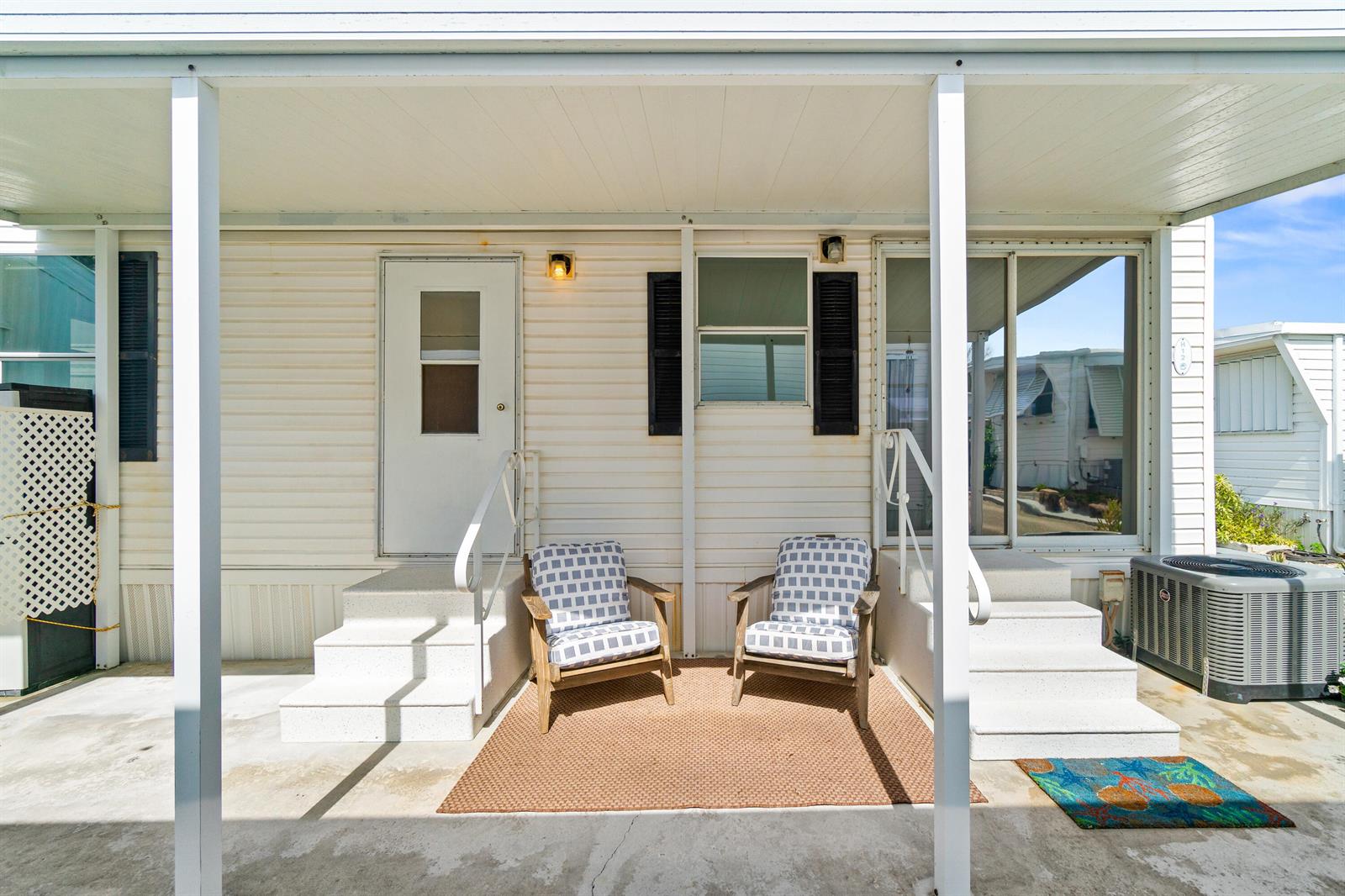 A covered patio features two chairs on a rug between a door and sliding glass doors — 12 Hibiscus Drive # H, Briny Breezes FL 33435, BRINY BREEZES, FL