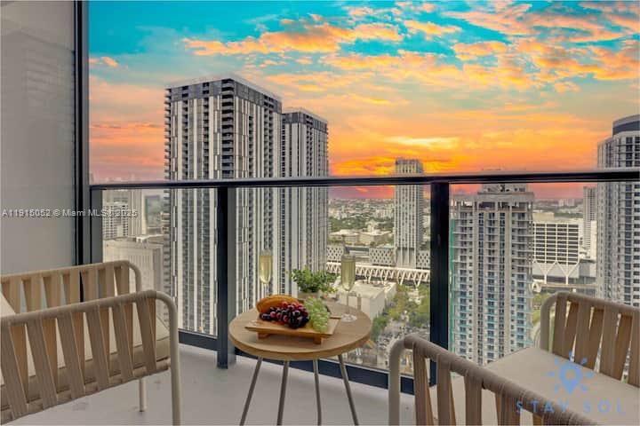 A balcony with two chairs and a small table overlooking a city skyline at sunset — 159 NE 6th St # 3004, Miami FL 33132, MIAMI, FL