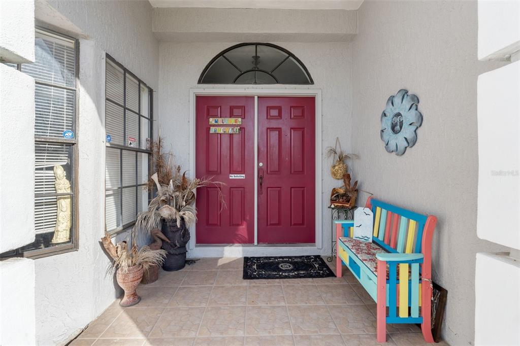 A covered front porch features a red double door, tiled flooring, and a colorful bench — 13732 TRIUMPH COURT, HUDSON, FL