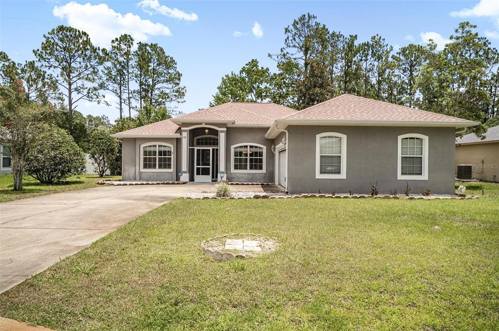A single-story gray house with a tan shingled roof, concrete driveway, and surrounding pine trees — 66 RAMBLEWOOD DRIVE, PALM COAST, FL