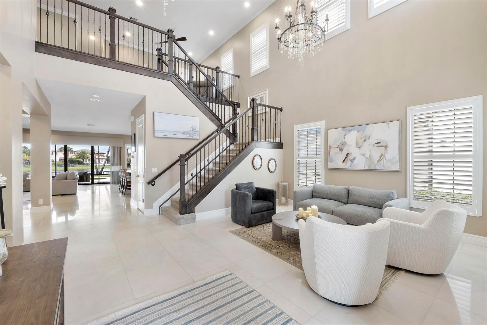 A high-ceilinged living room features a dark wood staircase, tile flooring, and a crystal chandelier — 11525 NW 81st Place, Parkland FL 33076, PARKLAND, FL
