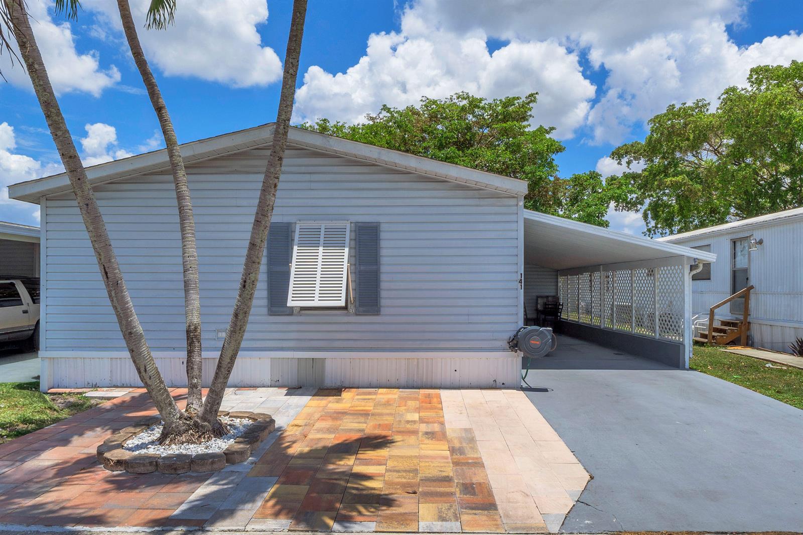 A light blue mobile home features a paved front patio, palm tree, and attached carport — 141 Plantation Boulevard, Lake Worth FL 33467, LAKE WORTH, FL