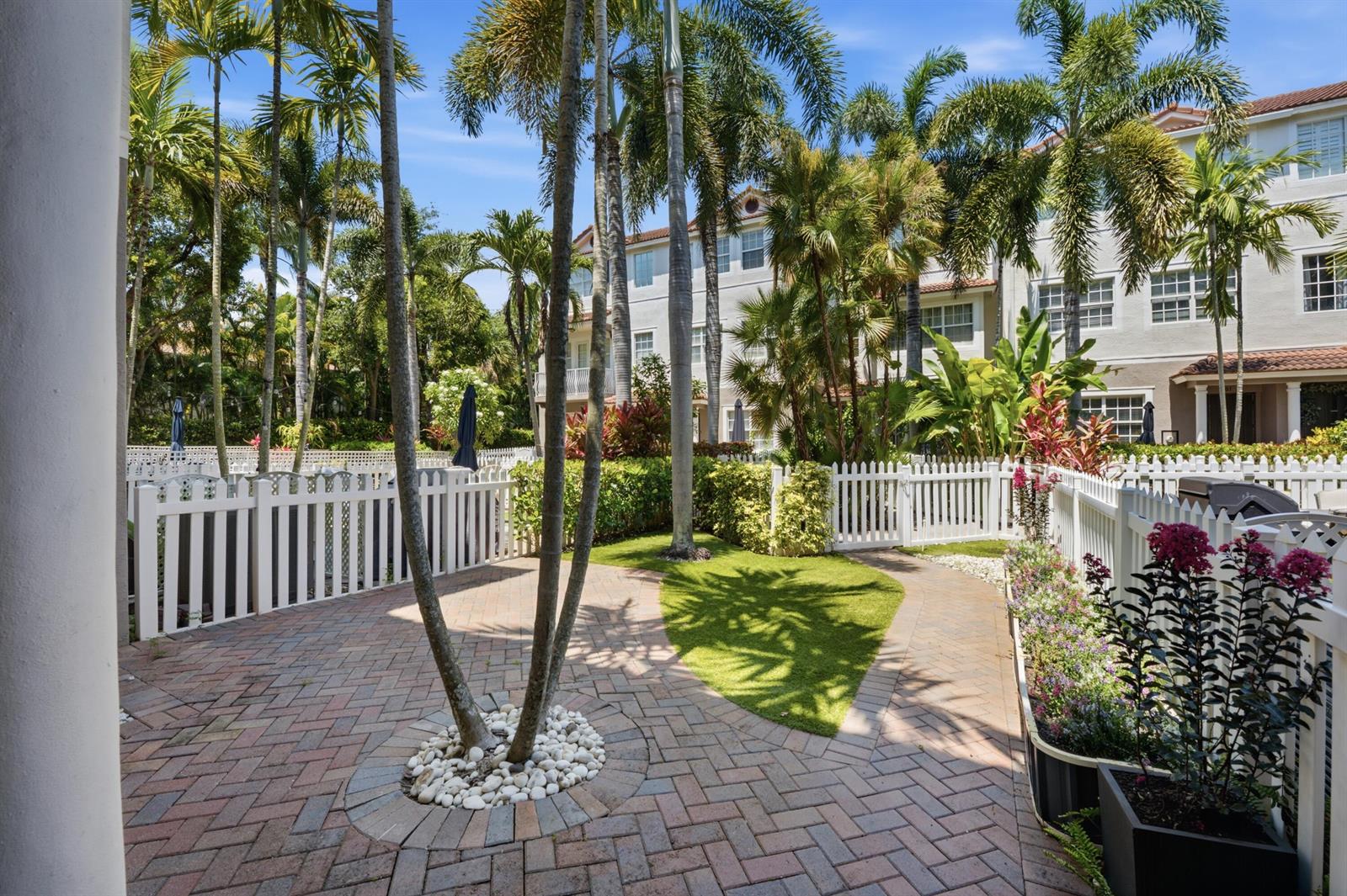 A brick patio features a palm tree, a small patch of grass, and a white fence — 107 Ocean Cay Way, Hypoluxo FL 33462, HYPOLUXO, FL