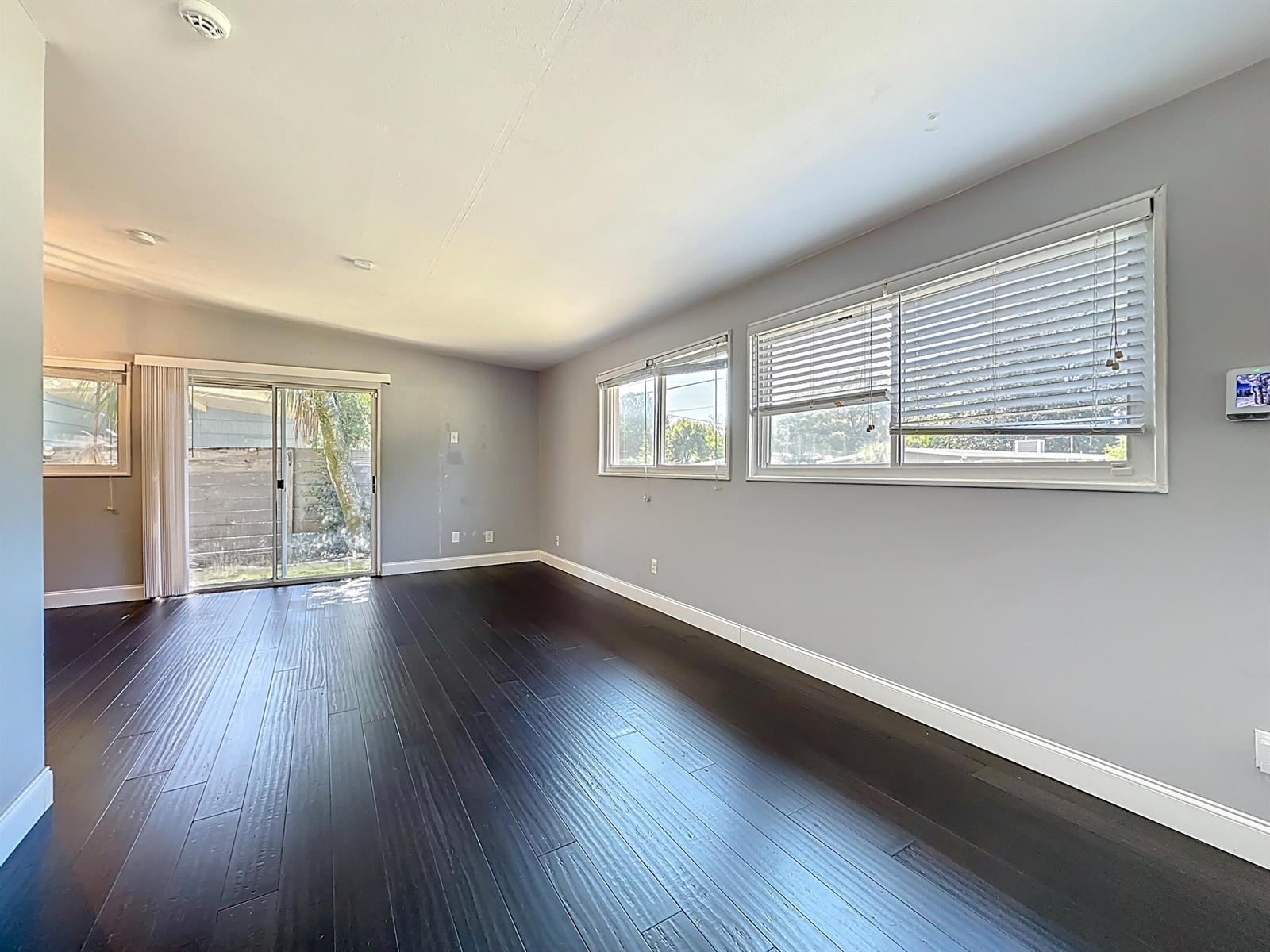 An empty room features dark wood flooring, grey walls, large windows, and a sliding glass door — 7615 Mayapple Road, Jacksonville FL 32211, JACKSONVILLE, FL