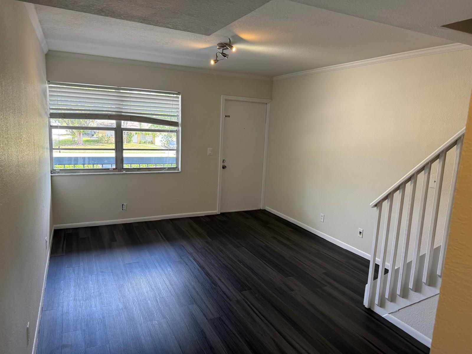 An empty living room features dark wood-look flooring, a window with blinds, and a staircase — 9500 SW 3rd Street # 222, Boca Raton FL 33428, BOCA RATON, FL