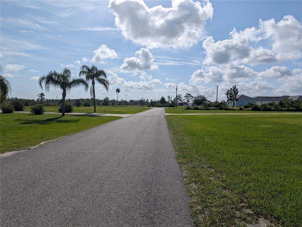 A paved road stretches through a grassy, undeveloped residential lot under a cloudy blue sky — 7 POINCIANA DRIVE, INDIAN LAKE ESTATES, FL