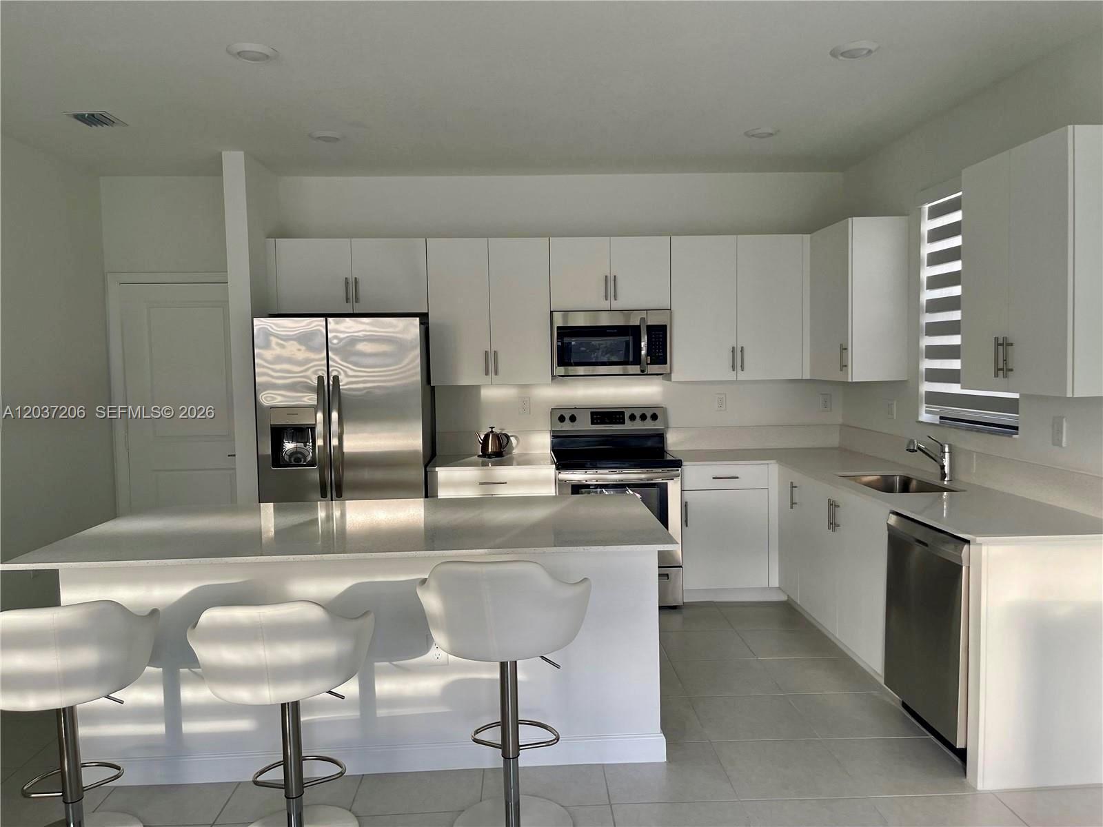 A modern kitchen features white cabinetry, stainless steel appliances, a quartz island, and bar stools — 22960 SW 130th Ave, Miami FL 33170, MIAMI, FL