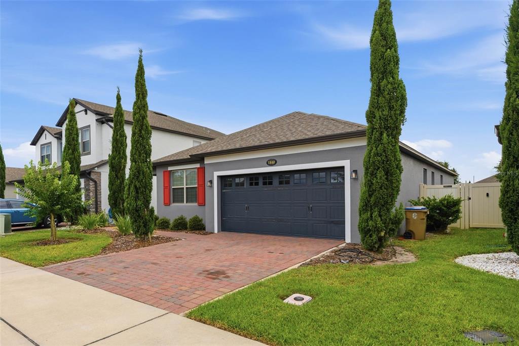 A gray single-story house features a brick driveway, a dark garage door, and tall cypress trees — 1840 NAPOLI DRIVE, ST CLOUD, FL