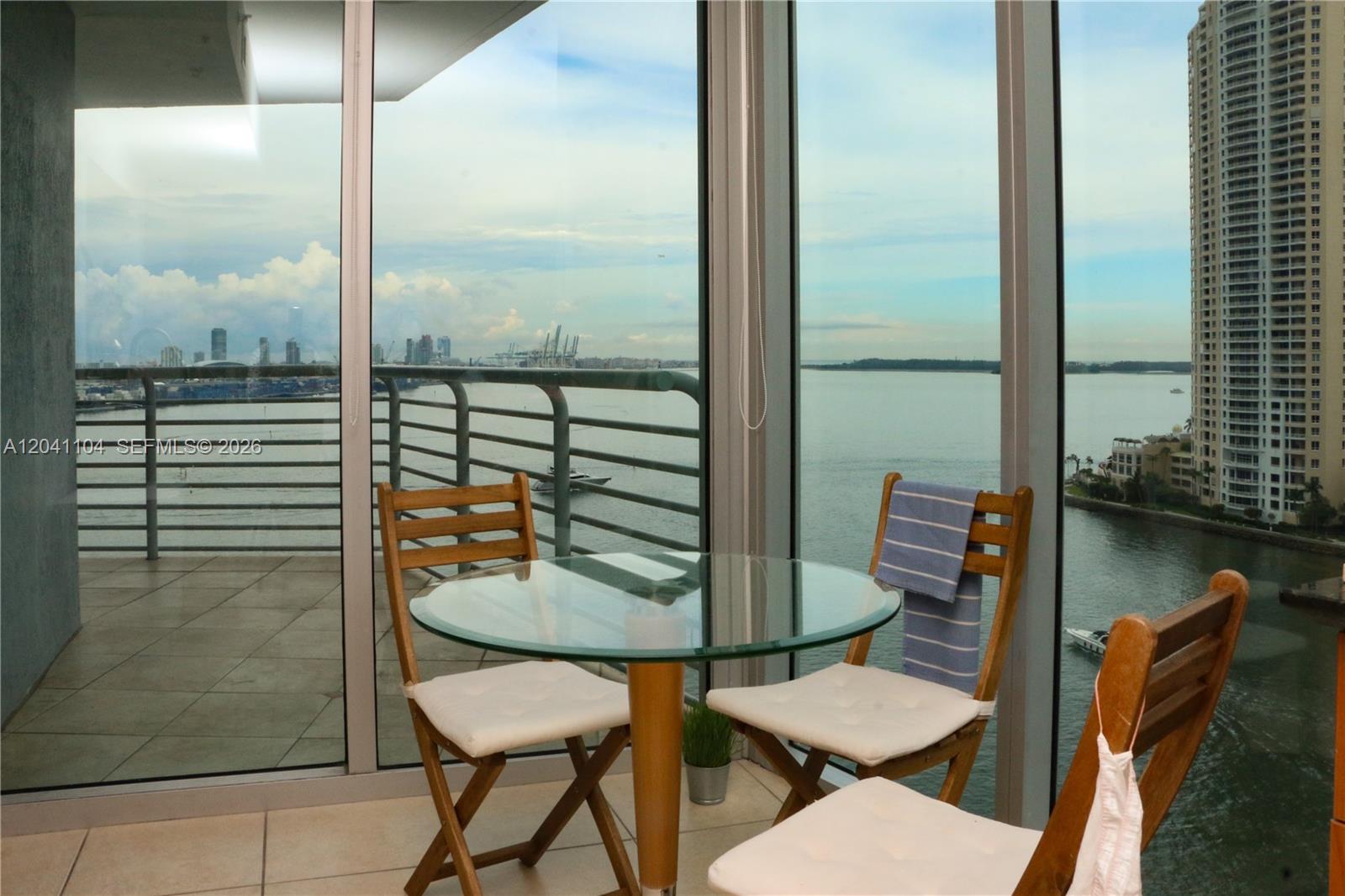 A glass-topped table and three wooden chairs sit on a balcony overlooking the water — 325 S Biscayne Blvd # 1523, Miami FL 33131, MIAMI, FL