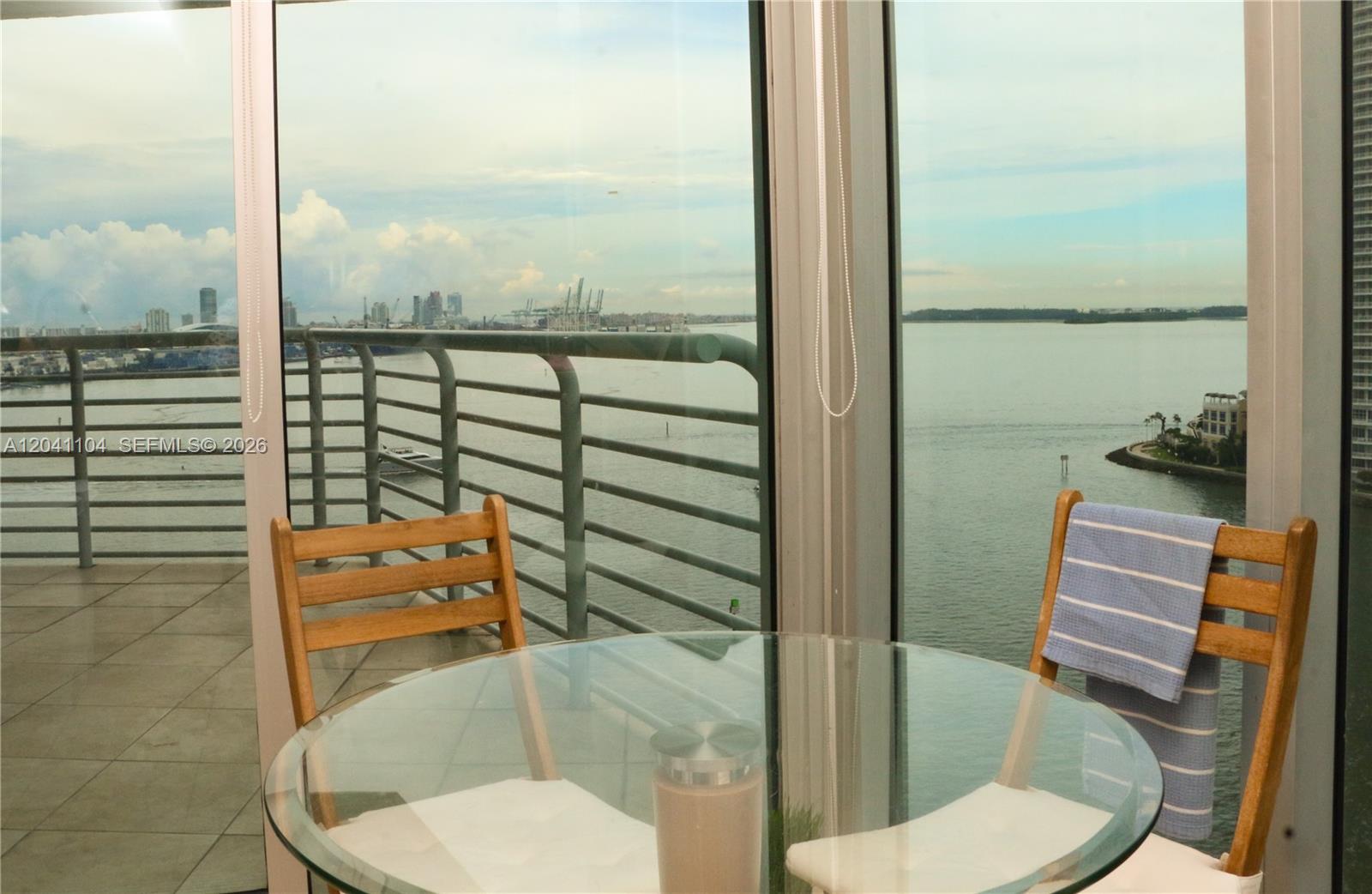 A glass-topped table and two wooden chairs sit on a balcony overlooking the water — 325 S Biscayne Blvd # 1523, Miami FL 33131, MIAMI, FL