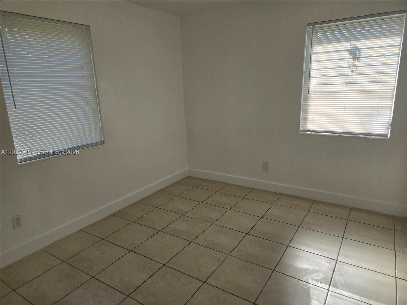 An empty room features white walls, tan tile flooring, and two windows with blinds — 408 NW 8th St # 408, Hallandale Beach FL 33009, HALLANDALE BEACH, FL