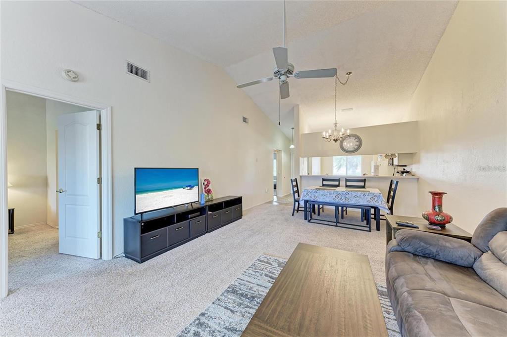 A living room with vaulted ceilings, carpeted floors, a television console, and an adjacent dining area — 4366 MADEIRA COURT UNIT 3356, SARASOTA, FL