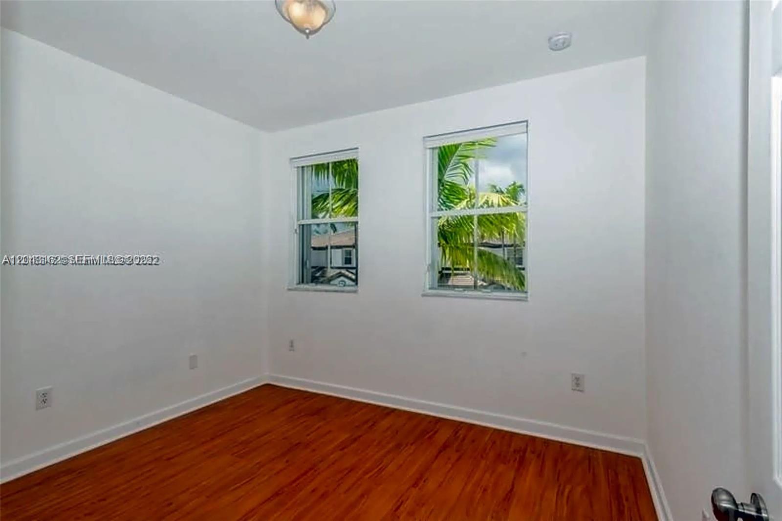 An empty bedroom features white walls, wood-look flooring, and two windows overlooking palm trees — 11497 NW 78th Ter, Doral FL 33178, DORAL, FL