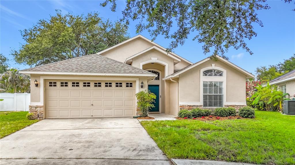 A single-story suburban house with a tan stucco exterior, a two-car garage, and a lawn — 7016 JAMESTOWN MANOR DRIVE, RIVERVIEW, FL