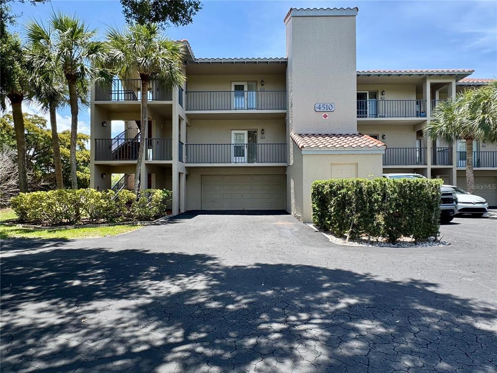 A three-story apartment building features a ground-level garage, balconies, and a paved driveway — 4510 EL CONQUISTADOR PARKWAY UNIT 204, BRADENTON, FL