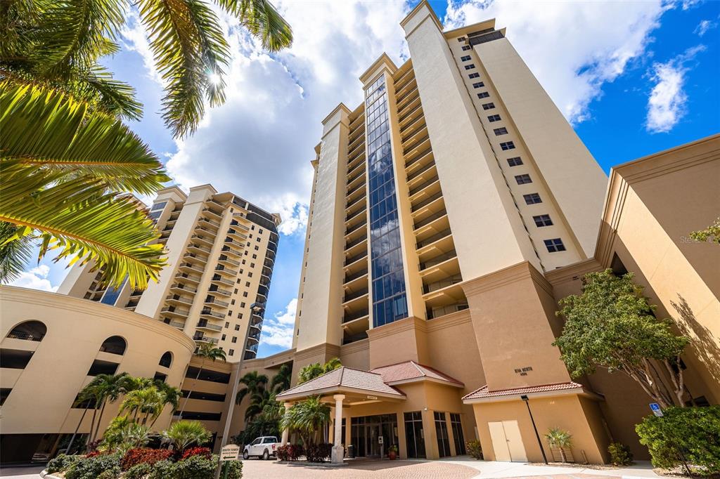 A low-angle exterior view of two tall, beige residential towers with glass balconies and palm trees — 14300 RIVA DEL LAGO DRIVE UNIT 2104, FORT MYERS, FL