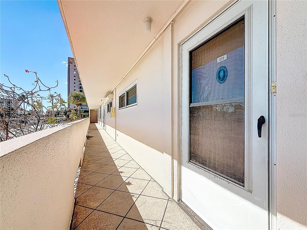 An outdoor walkway with tiled flooring leads past a white door marked 232 — 7401 BAY ISLAND DRIVE S UNIT 232, SOUTH PASADENA, FL