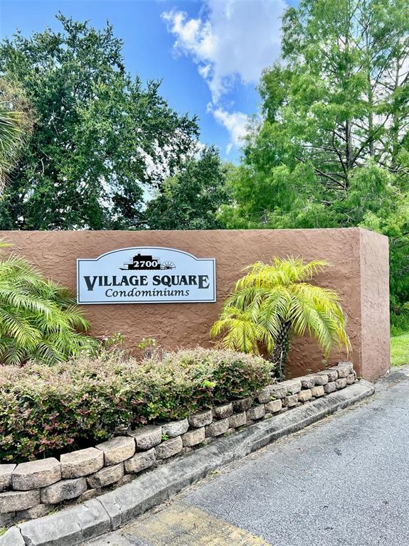 A brown stucco wall features a sign for Village Square Condominiums with landscaping and trees — 6065 VILLAGE CIRCLE UNIT 6065, ORLANDO, FL
